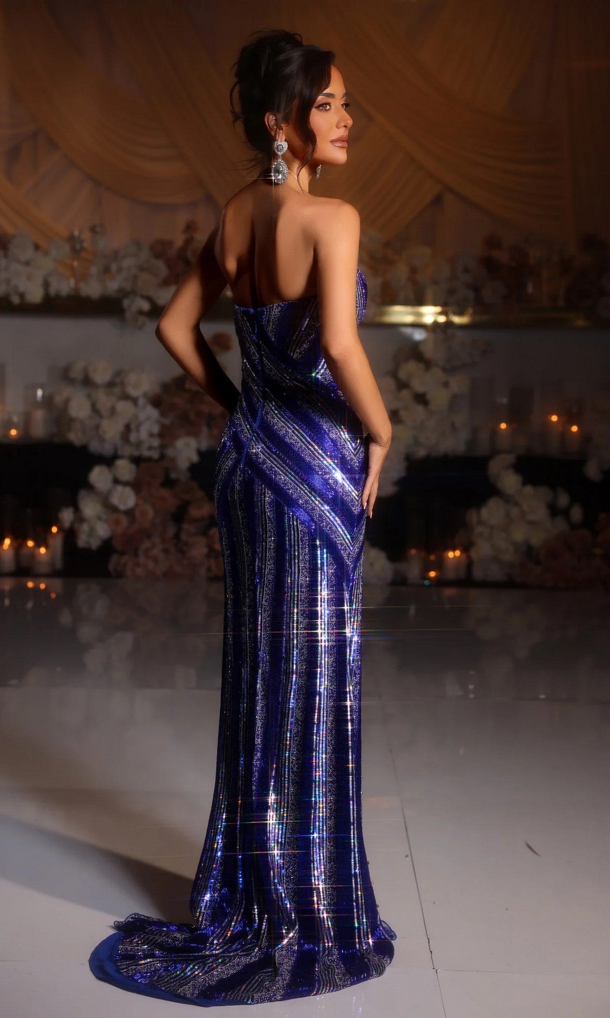 A woman wearing the Portia and Scarlett PS25449C long formal dress poses indoors with her back to the camera, surrounded by candles and floral arrangements.